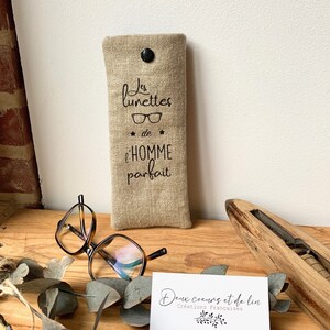 May include: A beige linen glasses case with black text that reads "Les lunettes de l'homme parfait" (The glasses of the perfect man). The case is sitting on a wooden surface with a pair of tortoiseshell glasses and a business card that reads "Deux coeurs et de lin Creations Francaises".