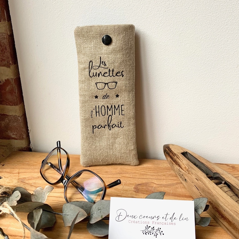 May include: A beige linen glasses case with black text that reads "Les lunettes de l'homme parfait" (The glasses of the perfect man). The case is sitting on a wooden surface with a pair of tortoiseshell glasses and a business card that reads "Deux coeurs et de lin Creations Francaises".