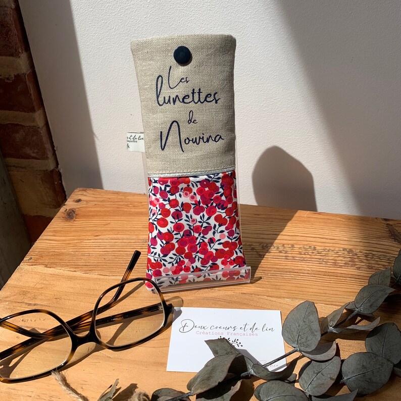 May include: A beige linen glasses case with the text "Les lunettes de Nouria" embroidered in blue. The case has a red and white floral lining and a blue button closure. The case is sitting on a wooden surface with a business card that reads "Deux coeurs et de lin Creations Franco-japonaises".