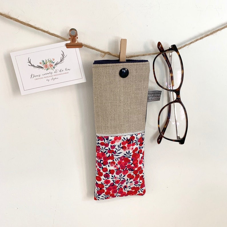 May include: A beige and red floral glasses case with a navy blue trim and a button closure. The case is hanging from a string with a pair of glasses next to it.