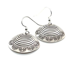May include: A pair of silver earrings with a shell design. Each earring has horizontal lines and stylized face motifs around the edge. The earrings have a hook closure. The earrings are displayed against a white background.