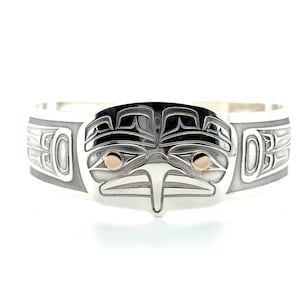 Peut inclure: Bracelet manchette argenté avec un motif central de visage d'oiseau stylisé. Les yeux de l'oiseau sont mis en valeur par des détails dorés. Le bracelet présente des motifs gravés sur les côtés, art amérindien.