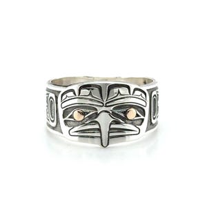 Peut inclure: Une bague en argent avec un motif amérindien détaillé. La bague présente un visage central en forme d'oiseau avec des accents dorés pour les yeux. La bande est gravée de motifs tribaux supplémentaires.