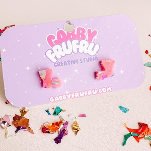 Pode incluir: Brincos rosa e multicoloridos em forma de bota em um cartão lavanda com o texto "Gabby FruFru Creative Studio" e "gabbyfrufru.com". Os brincos são cercados por confetes coloridos.