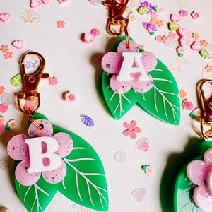 May include: Keychains with a floral design. Each keychain features a green leaf, a pink flower with glitter, and a white letter. The keychains have gold-colored clasps. Decorative floral embellishments are scattered around the keychains.