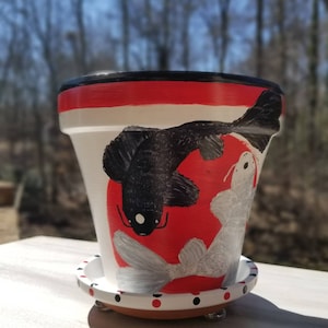 May include: White ceramic planter with a red, black, and white design of two koi fish. The planter has a matching saucer.