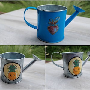 May include: Three small metal watering cans. One is blue with a heart design and the other two are silver with a pineapple design.