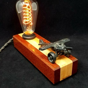 May include: A wooden lamp base with a red and natural wood finish features a small cast iron airplane and a vintage Edison bulb.