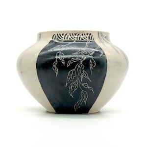 May include: A white ceramic vase with a black and white design of leaves and branches. The vase has a rounded shape and a wide opening.