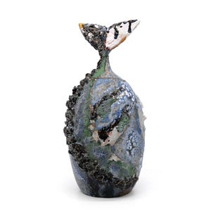 Op de afbeelding: Een keramische sculptuur van een walvis met een getextureerd, veelkleurig lichaam. De sculptuur heeft een staartvin met zwarte, witte en gele accenten. Het lichaam is voornamelijk blauw, groen en grijs met een ruw, getextureerd oppervlak.