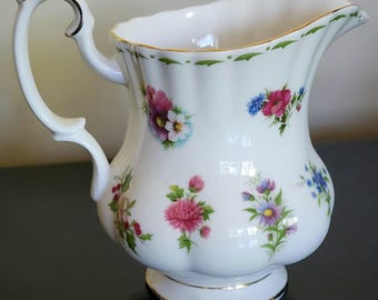 Puede incluir: Jarra de cerámica blanca con asa curva y estampado floral. La jarra presenta un borde festoneado con ribete verde y detalles dorados. El diseño floral incluye flores rosas, moradas y azules.