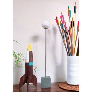 May include: A white ceramic sphere on a silver stand, a wooden rocket ship with a yellow tip and blue and white stripes, and a white ceramic vase filled with colorful arrows.