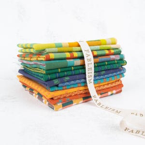 May include: A stack of colorful, patterned fabric squares in shades of yellow, green, blue, and orange, bound with a cream-colored ribbon printed with the word "FABLEISM". The fabrics have a woven texture and are neatly arranged.