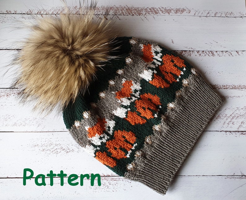 K&ouml;nnte beinhalten: Eine gestrickte Winterm&uuml;tze mit einem braunen Fellbommel. Die M&uuml;tze hat ein Muster aus orangefarbenen F&uuml;chsen, wei&szlig;en Akzenten und einem dunkelgr&uuml;nen und grauen Farbschema. Das Wort "Pattern" ist gr&uuml;n sichtbar.