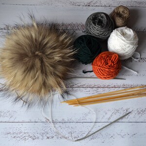 K&ouml;nnte beinhalten: Ein flauschiger, brauner Fellbommel liegt neben mehreren Wollkn&auml;ueln in Wei&szlig;, Orange, Gr&uuml;n, Grau und Braun. Stricknadeln und eine Rundstricknadel sind ebenfalls vorhanden, was auf ein Bastelthema hindeutet.