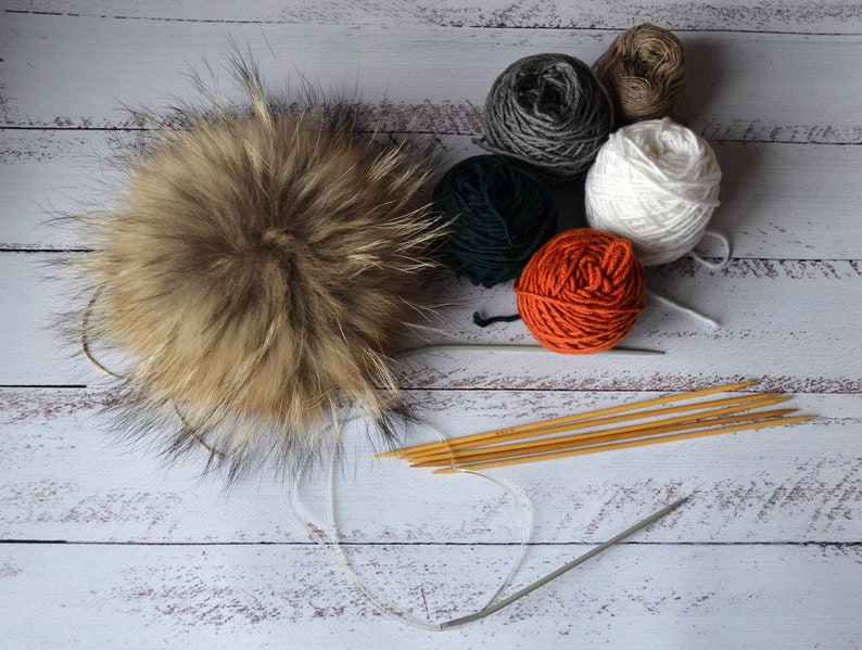 K&ouml;nnte beinhalten: Ein flauschiger, brauner Fellbommel liegt neben mehreren Wollkn&auml;ueln in Wei&szlig;, Orange, Gr&uuml;n, Grau und Braun. Stricknadeln und eine Rundstricknadel sind ebenfalls vorhanden, was auf ein Bastelthema hindeutet.