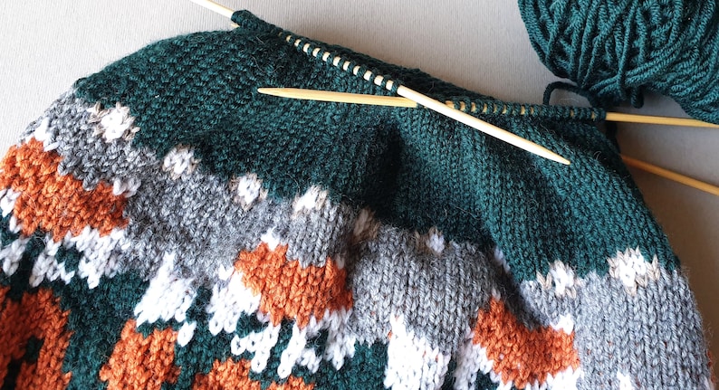 K&ouml;nnte beinhalten: Ein teilweise fertiggestellter Strickpullover in Gr&uuml;n-, Grau-, Wei&szlig;- und Oranget&ouml;nen. Der Pullover hat ein geometrisches Muster und ist in Arbeit, mit h&ouml;lzernen Stricknadeln und einem Kn&auml;uel Garn sichtbar.