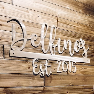 May include: A silver metal sign with the name "Delbino's Est 2015" on a wood wall. The sign is attached to the wall with a metal bar. The text "#onelovemetals onelovemetals.etsy.com" is visible below the sign.