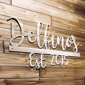 May include: A silver metal sign with the name "Delbino's" and the text "Est 2015" on a wood wall. The sign is attached to the wall with a metal bar. The text "#onelovemetals onelovemetals.etsy.com" is visible at the bottom of the image.