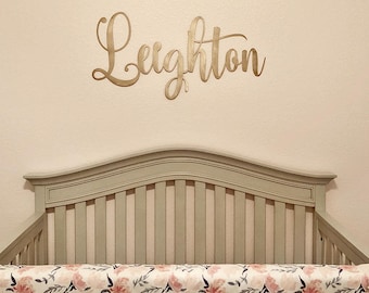 May include: A gold metal name sign with the name "Leighton" in cursive script, above a white crib with a pink and floral patterned crib skirt. The text "#onelovemetals onelovemetals.etsy.com" is printed below the crib.