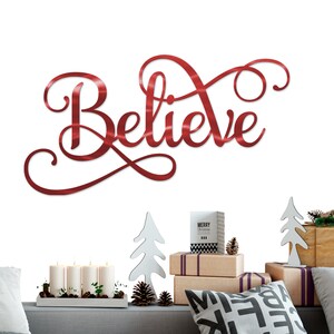 May include: Red metal Believe sign with a cursive font. The sign is hanging on a white wall above a couch with a white throw pillow and a black and white patterned throw pillow. There are also small white Christmas trees and a box that says "Merry Christmas" on the couch.