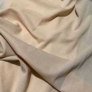 May include: A close-up of a beige fabric with a smooth, soft texture. The fabric is draped and folded, creating a sense of movement and flow.