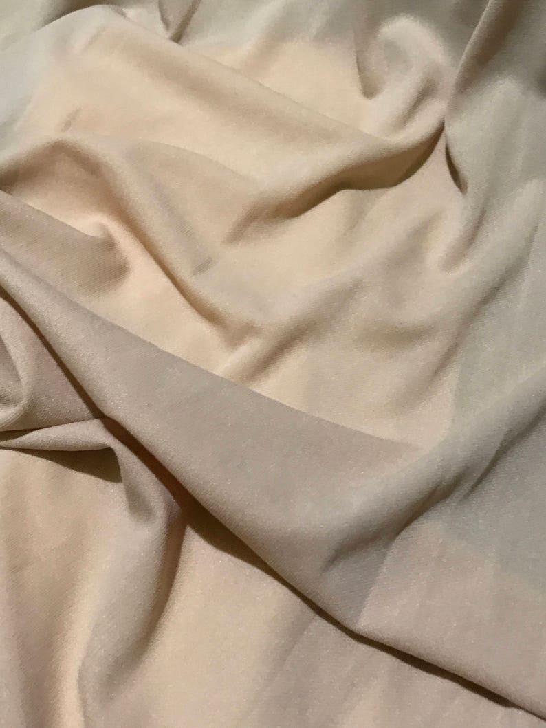 May include: A close-up of a beige fabric with a smooth, soft texture. The fabric is draped and folded, creating a sense of movement and flow.