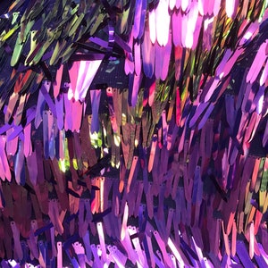 May include: Close-up of a shimmering fabric with a multitude of iridescent, elongated sequins in shades of purple, pink, and gold. The sequins are arranged in a dense, overlapping pattern, creating a textured, reflective surface. The background is a dark mesh.
