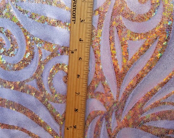 May include: Close-up of a white fabric with a gold and iridescent swirl pattern. A wooden ruler is placed on the fabric to show the scale. The ruler is marked with inches.