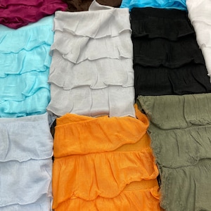 K&ouml;nnte beinhalten: Eine Sammlung von zehn Stoffstreifen mit gekr&auml;uselten Kanten in verschiedenen Farben, darunter hellblau, wei&szlig;, orange, gr&uuml;n, schwarz, braun, rosa und lila. Die Stoffstreifen sind in einem vertikalen Stapel angeordnet.