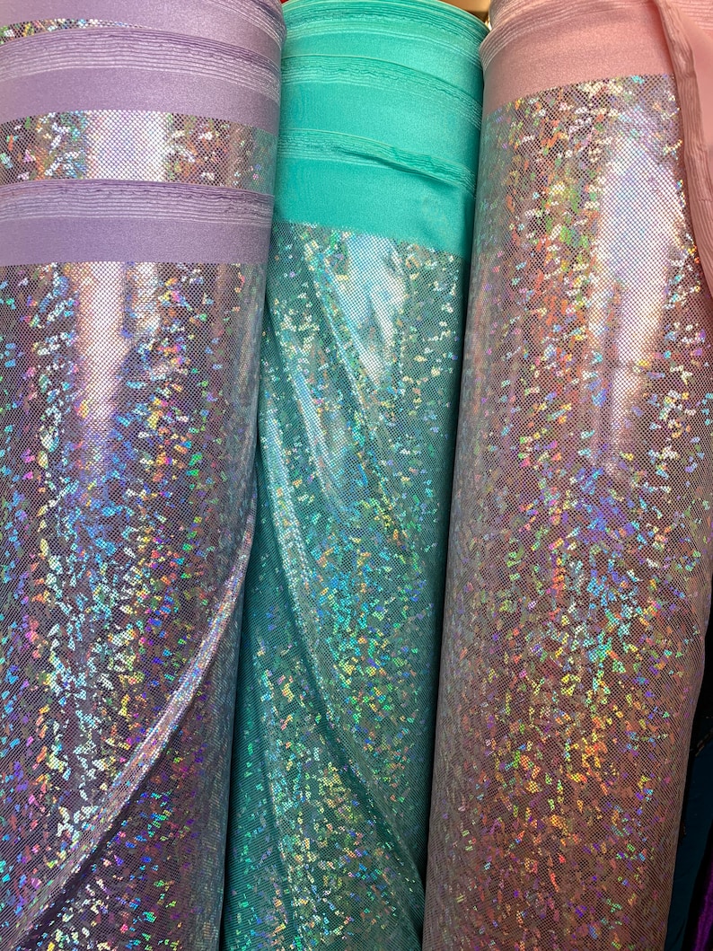 May include: Three rolls of iridescent fabric in lavender, teal, and pink. The fabric has a holographic finish with a subtle sparkle.