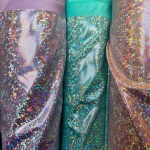 May include: Three rolls of iridescent fabric with a holographic finish. The fabric is a light purple, teal, and a light brown color.