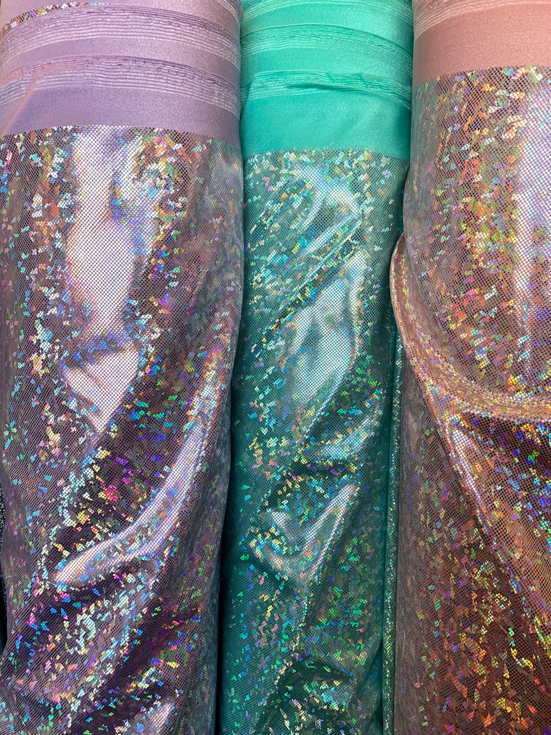 May include: Three rolls of iridescent fabric with a holographic finish. The fabric is a light purple, teal, and a light brown color.