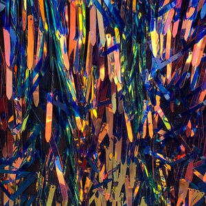 May include: A close-up of iridescent blue and gold fringe fabric. The fringe is made of thin, flat strips of fabric that are sewn together to create a flowing, shimmering effect.