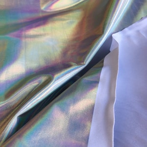 May include: Close-up of iridescent fabric with a holographic finish. The fabric is folded over itself, revealing a white backing.