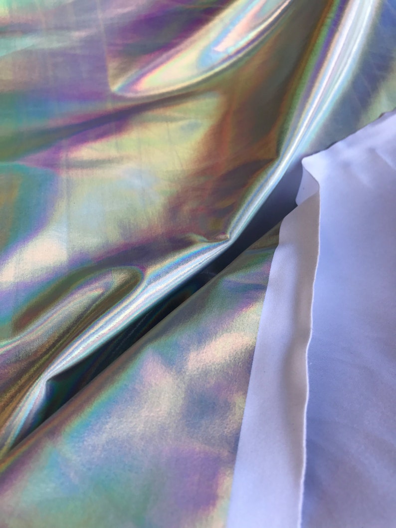 May include: Close-up of iridescent fabric with a holographic finish. The fabric is folded over itself, revealing a white backing.