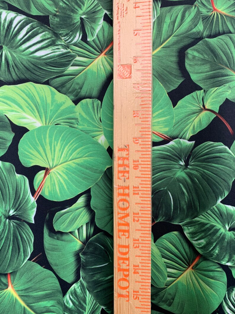 May include: Close-up of a black fabric with a green tropical leaf print. A wooden ruler with the text "THE HOME DEPOT" is placed next to the fabric.
