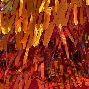 May include: Close-up of a red fabric with gold fringe trim. The fringe is made of thin, metallic strips that are sewn together in a staggered pattern.