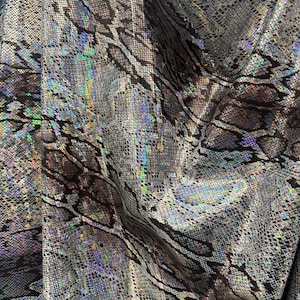 Peut inclure: Gros plan sur un tissu à motif peau de serpent. Le tissu a un éclat métallique avec des reflets irisés bleus, verts et violets. La couleur de base est un mélange de gris et de noir, créant un aspect texturé.