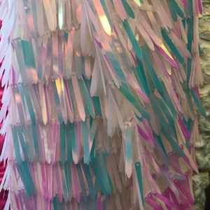 May include: Close-up of iridescent sequins in white, blue, and purple shades, arranged in a layered, fringe-like pattern. The sequins are sewn onto a sheer, white mesh fabric.