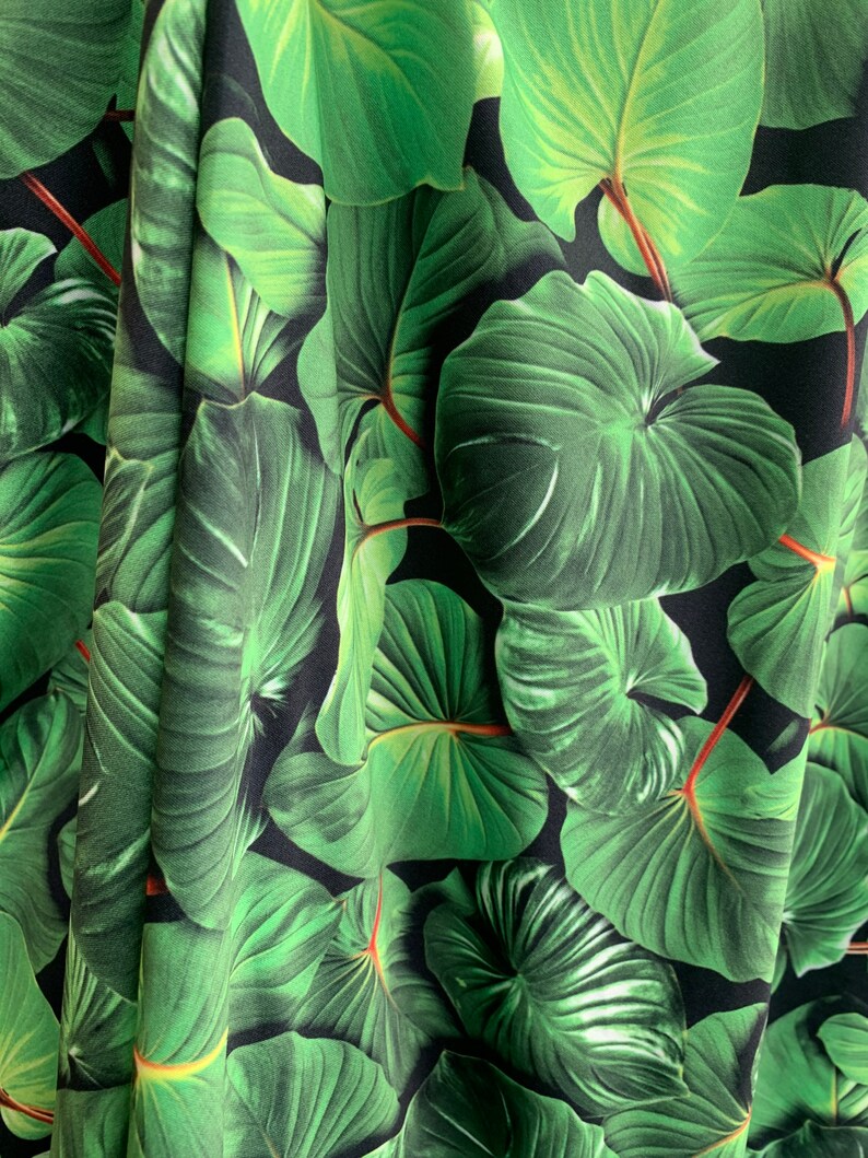 May include: Close-up of a fabric with a green tropical leaf pattern on a black background. The leaves are large and have a realistic texture.