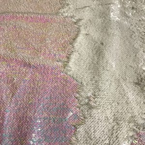 May include: A close-up of iridescent fabric with a two-tone design. One side is a light pink and blue colour, while the other side is a silver colour. The fabric is made of sequins.
