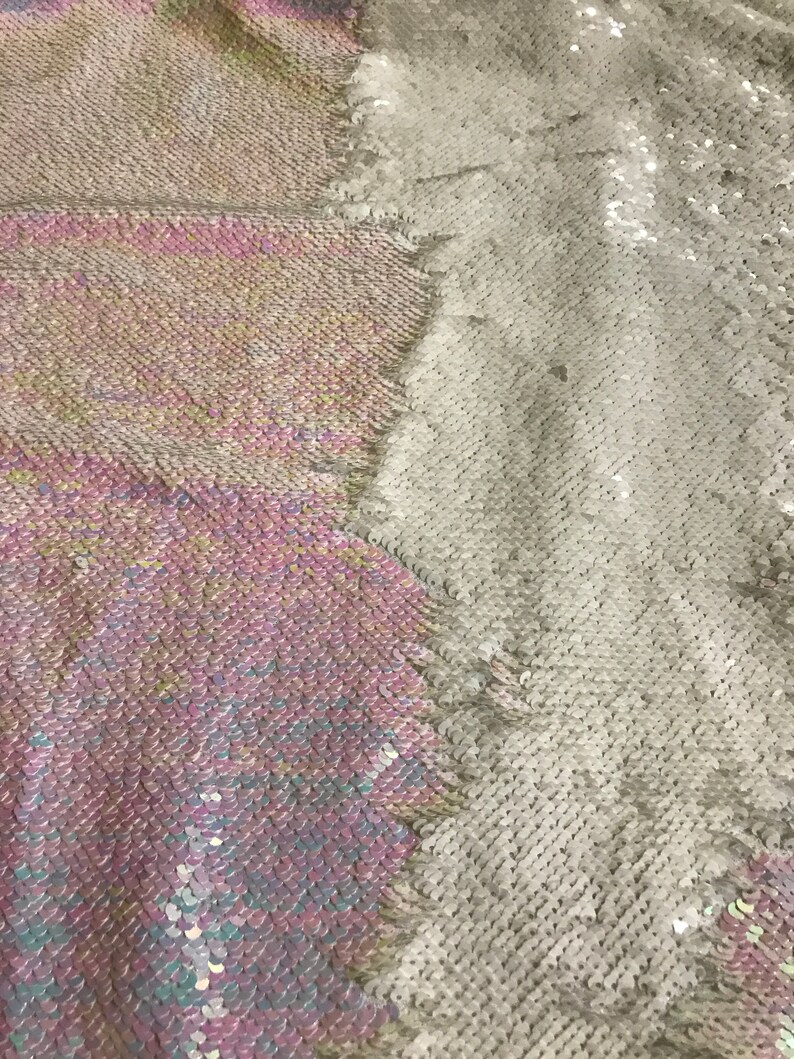 May include: A close-up of iridescent fabric with a two-tone design. One side is a light pink and blue colour, while the other side is a silver colour. The fabric is made of sequins.