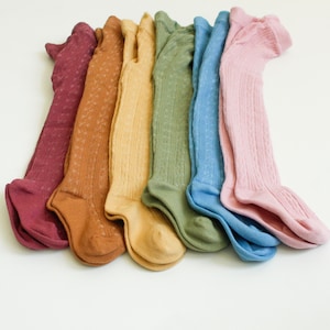 May include: A set of seven pairs of colorful tights with a textured knit pattern. The tights are arranged in a rainbow order, starting with maroon and ending with light pink.