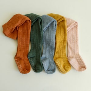 May include: Five pairs of colorful tights with a cable knit pattern. The tights are in shades of orange, green, blue, yellow, and pink. Each pair has a white heart pattern on the bottom.