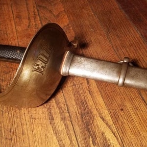 May include: A silver sword with a brass guard and a black handle. The guard is engraved with the word "E.U.P."