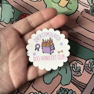 May include: A white, scalloped-edge sticker with the text "OVERSTIMULATED DOG HANDLERS CLUB" and a graphic of a purple dumpster on fire. The sticker also features a dog bone, paw prints, and a ribbon.