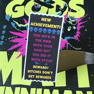 May include: A book cover with the title "GODS" in large, neon yellow letters. A sticker with the text "NEW ACHIEVEMENT!" and "YOU NO'D IN THE RING WITH YOUR DOGI BUT YOU DID IT WITH STYLE!" is placed on the book.