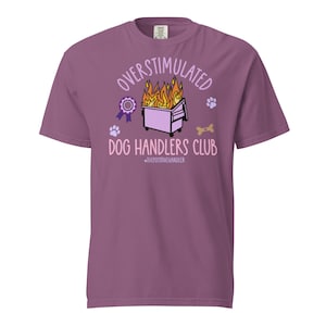 May include: A mauve short-sleeve t-shirt featuring a graphic of a dumpster on fire. The text reads OVERSTIMULATED DOG HANDLERS CLUB and #THEPOSITIVEHANDLER. The design includes a purple award ribbon, two light purple paw prints, and a tan bone.