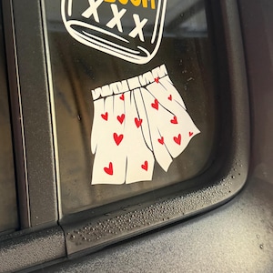 May include: A white vinyl decal of a pair of boxer shorts featuring a red heart pattern, positioned below a white and yellow decal of a jug with the text JUG O' BOOM XXX, both applied to a dark tinted vehicle window surface.